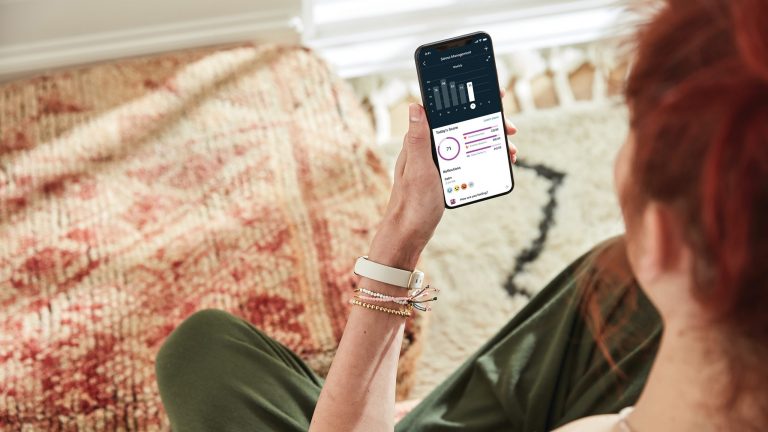Eine Person sitzt auf einem Teppich und schaut auf das Smartphone in ihrer Hand. Darauf wird gerade eine Statistik in der Fitbit-App angezeigt.