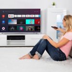 Eine Person sitzt auf einem Teppich vor einem Sofa und schaut zum Fernseher an der Wand, in der Hand eine Fernbedienung.