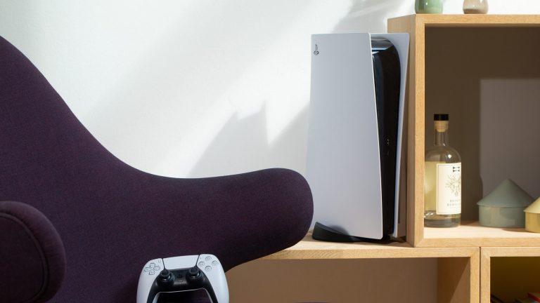 Der PS5 DualSense-Controller lehnt auf einem lila-farbenen Sessel. Im Hintergrund steht die PS5 in einem Regal.