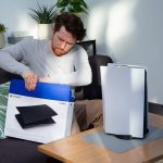 Eine Person sitzt auf einem Sofa vor einer PS5 und zieht schwarze Seitenteile f&uuml;r die Konsole aus einem Karton.