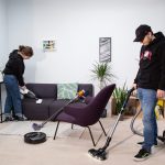 Zwei Personen saugen einen Raum mit Handstaubsauger und Bodenstaubsauger, auf dem Boden steht ein Saugroboter, an einem Sofa lehnt ein Stielsauger.