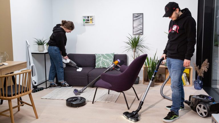 Zwei Personen saugen einen Raum mit Handstaubsauger und Bodenstaubsauger, auf dem Boden steht ein Saugroboter, an einem Sofa lehnt ein Stielsauger.