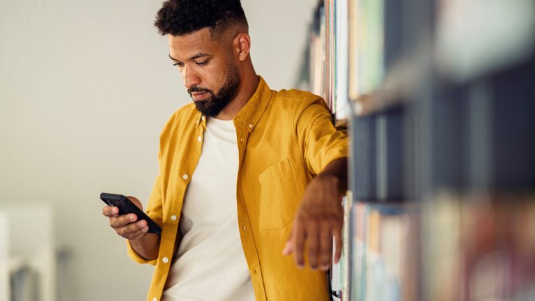 Eine Person lehnt in einer Bibliothek an ein Bücherregal und schaut auf ihr Smartphone.