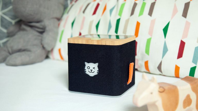 Eine schwarze Tigerbox Touch steht auf einem Sofa. Dahinter liegen ein farbenfrohes Kissen, ein Stoffnasshorn und eine Spielzeugkuh aus Holz.
