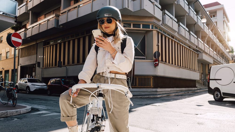 Eine Person steht mit einem Fahrrad auf einer Straße und schaut auf ihr Smartphone.