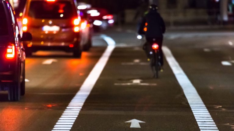 Auf einer Straße im Stadtverkehr steht eine Schlange von Autos mit leuchtendem Bremslicht. Daneben befindet sich ein Fahrradweg, auf dem eine Person mit einem Fahrrad fährt.
