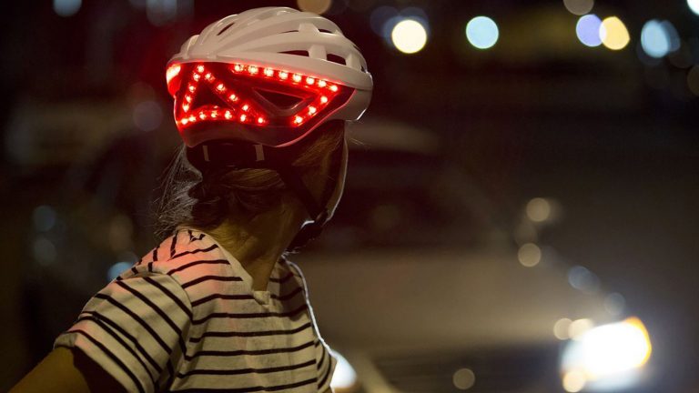 Eine Person trägt den Fahrradhelm Kickstart von Lumos. Sie wirft einen Blick über ihre Schulter, sodass die leuchtenden LEDs an der Rückseite des Helms zu sehen sind.