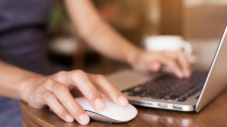 Eine Person steht vor einem Laptop und bedient diesen mit einer Maus.