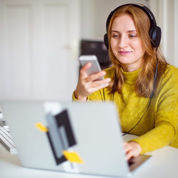 Eine Person sitzt vor einem Laptop und schaut auf ein Smartphone in ihrer Hand.