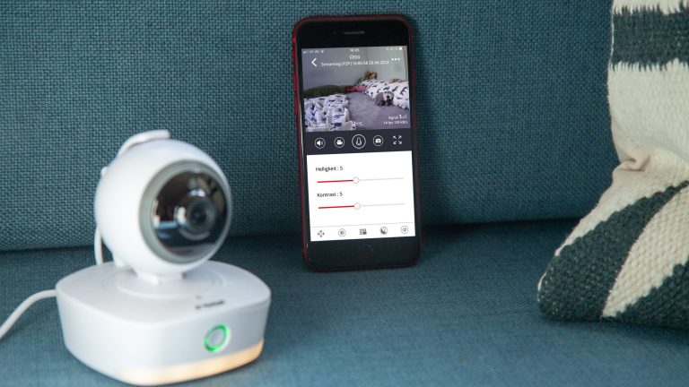 Eine Reer IP Babycam Move steht neben einem Smartphone auf einem Sofa. Auf dem Display sind diverse Einstellungsoptionen der Kamera zu erkennen.