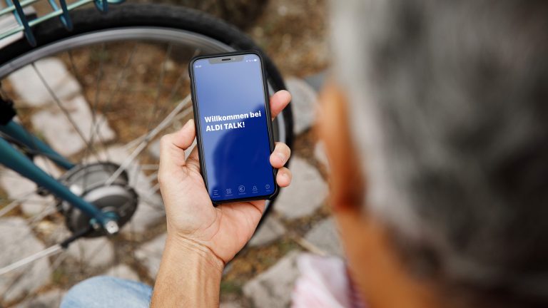 Eine Person blickt sitzend auf ihr Smartphone, auf dem die Aldi-Talk-App startet.