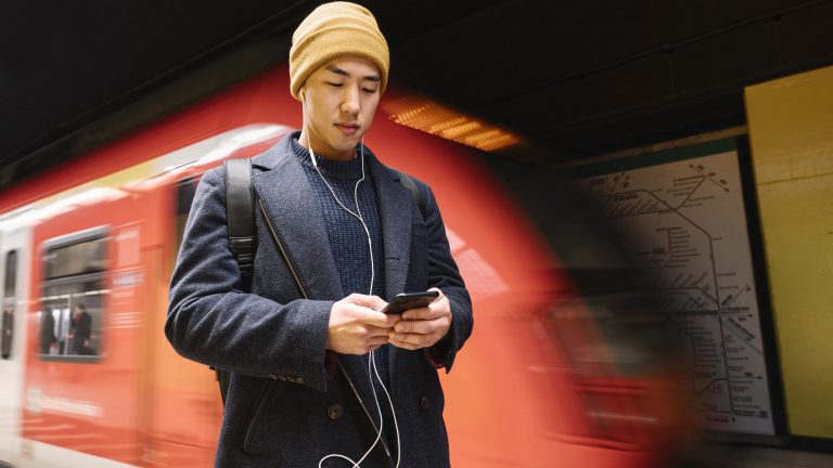 Eine Person steht auf dem Bahnsteig vor einer einfahrenden U-Bahn und schaut auf ihr Smartphone.
