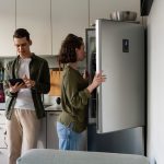 Ein Paar steht in der Küche, eine Person schaut auf ein Tablet, das sie in der Hand hält, die andere schaut in den geöffneten Kühlschrank.