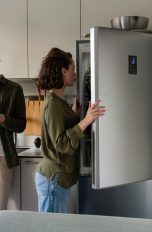 Ein Paar steht in der Küche, eine Person schaut auf ein Tablet, das sie in der Hand hält, die andere schaut in den geöffneten Kühlschrank.