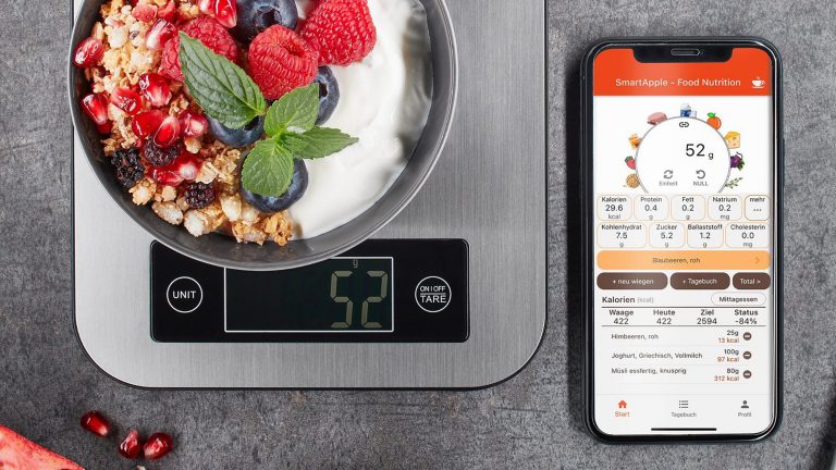 Auf einer K&uuml;chenwaage steht eine Sch&uuml;ssel mit Fr&uuml;chten, M&uuml;sli und Joghurt, daneben liegt ein Smartphone, auf dem eine App die N&auml;hrwerte der Lebensmittel anzeigt.