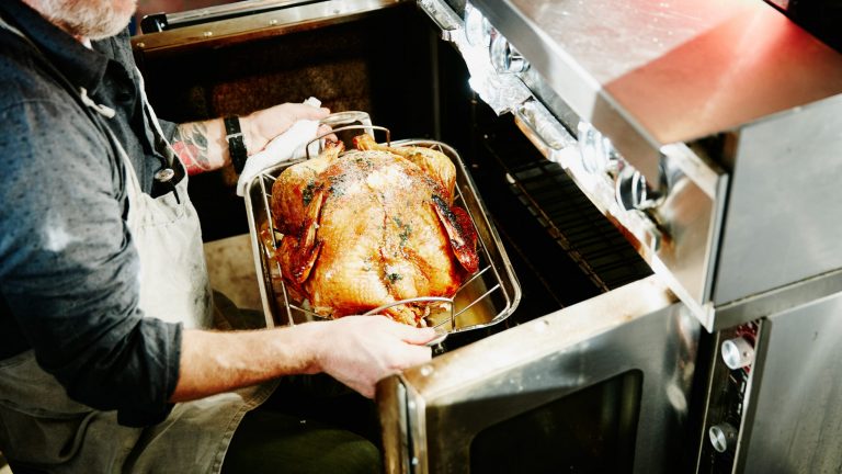 Eine Person kniet vor einem Backofen und holt ein Backh&auml;hnchen heraus.