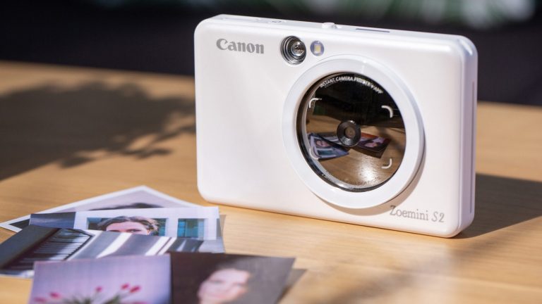 Eine Canon Zoemini S2 von vorne, auf der der spiegelnde Bereich um das Objektiv gut zu erkennen ist. Vor der Kamera liegen einige Ausdrucke.