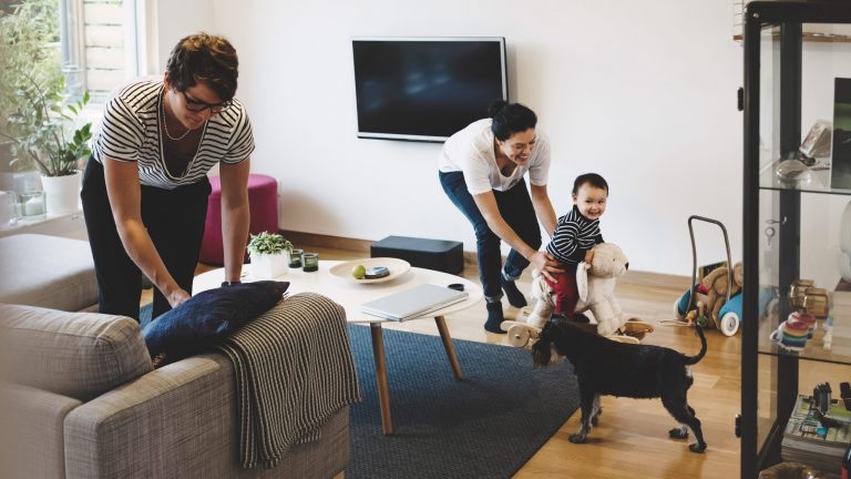 In einem Wohnzimmer mit Fernseher an der Wand befinden sich zwei Erwachsene, ein Kleinkind und ein Hund.