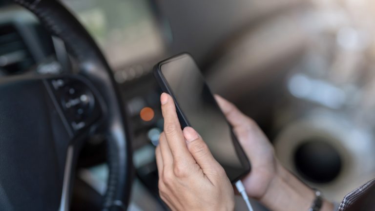 Eine Person sitzt auf dem Gfahrersitz eines Autos und hät ein Smartphone in den Händen, dass per USB-Kabel mit dem Autoradio verbunden ist.