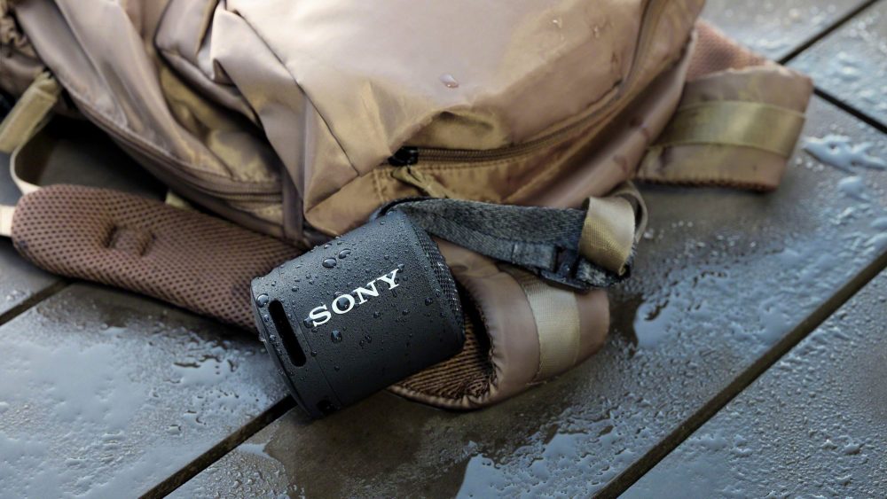 Ein schwarzer Sony SRS-XB13 hängt an einem nassen Rucksack.