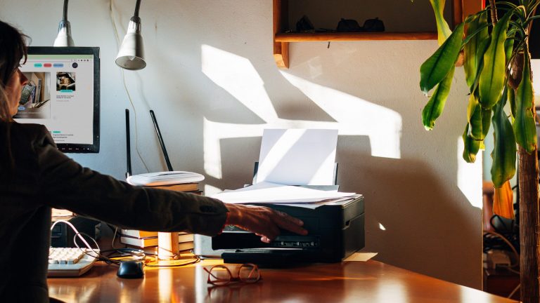 Eine Person sitzt an einem Schreibtisch und betätigt eine Taste am Drucker, der neben ihr steht.