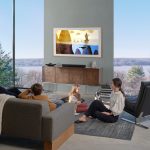 Zwei Eltern sitzen mit ihren beiden Kindern im Wohnzimmer vor einem Fernseher, der an der Wand hängt.