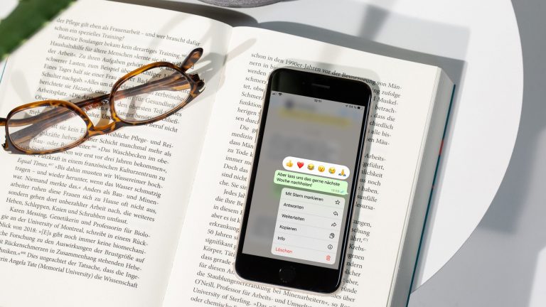 Ein Smartphone liegt auf einer Buchseite. Darauf zu sehen sind die Nachrichten-Optionen von WhatsApp.