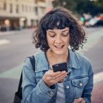 Eine Person steht an einer Stra&szlig;e und spricht in ihr Smartphone, das sie vor sich h&auml;lt.
