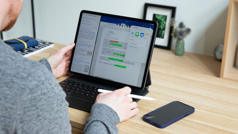 Eine Person sitzt vor einem iPad Pro, das in einem Magic Keyboard steckt. In der Hand hält die Person einen Apple Pencil 2, daneben liegt auf dem Holztisch ein iPhone 12.