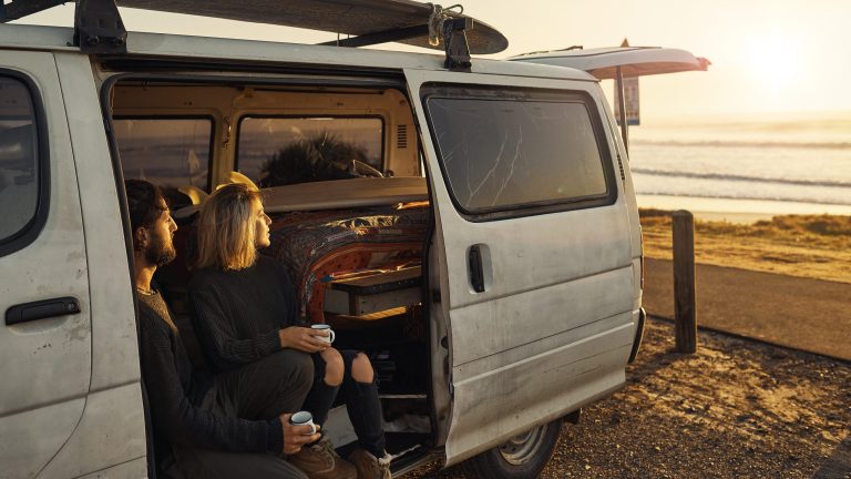Ein Paar sitzt in seinem Transporter bei geöffneter Schiebetür am Starnd und blickt Richtung Sonnenuntergang.