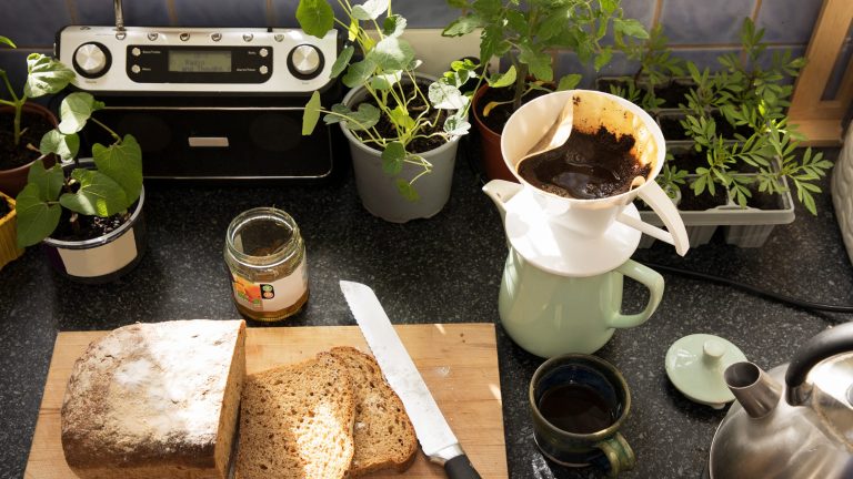 Ein DAB+-Radio steht in einer Küche auf der Arbeitsplatte, davor ein Holzbrett mit geschnittenem Brot, ein Glas Marmelade, eine Kaffeekanne mit Filter und eine Tasse.