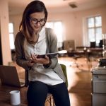 Eine Person hält ein iPad in der Hand. Im Hintergrund ist ein einem Büroraum ein Drucker zu sehen.