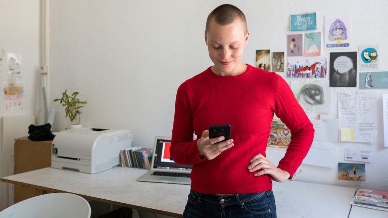 Eine Person steht mit einem iPhone in der Hand vor einem Schreibtisch, auf dem ein Drucker steht.