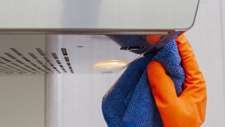 Eine Hand im orangefarbenen Gummihandschuh reinigt die Flächen einer Dunstabzugshaube mit einem Microfasertuch.