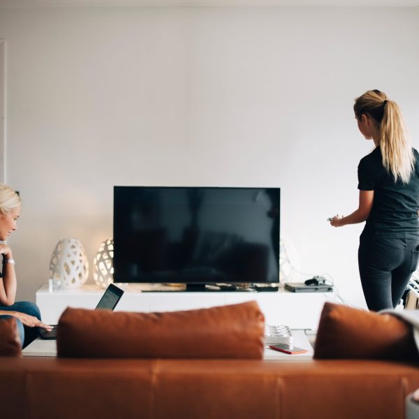 Zwei Personen vor einem Fernseher mit schwarzem Display, eine sitzt auf dem Sofa vor einem Laptop, die andere steht mit der Fernbedienung in der Hand.