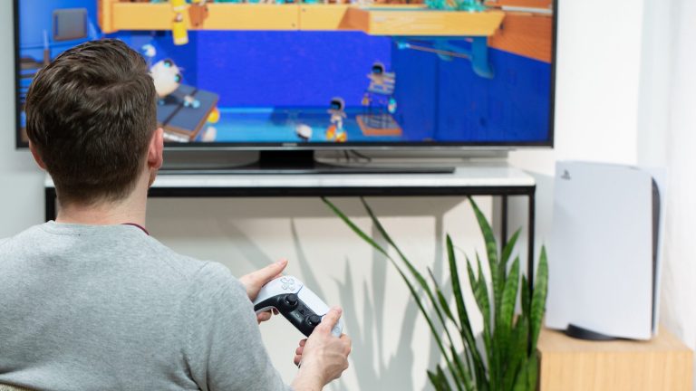 Eine Person sitzt vor einem Fernseher, neben dem eine PlayStation 5 steht. Sie hält einen Dualsense-Controller in der Hand.