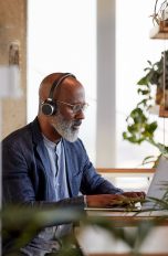 NAS einrichten Eine Person sitzt an einem Schreibtisch und tippt auf einem MacBook. Dabei trägt sie Kopfhörer. Im Vordergrund stehen einige Zimmerpflanzen.