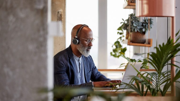 Eine Person sitzt an einem Schreibtisch und tippt auf einem MacBook. Dabei trägt sie Kopfhörer. Im Vordergrund stehen einige Zimmerpflanzen.