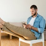 Eine Person sitzt auf einem Stuhl, hat die F&uuml;&szlig;e auf dem Tisch und schaut auf ihr Smartphone.