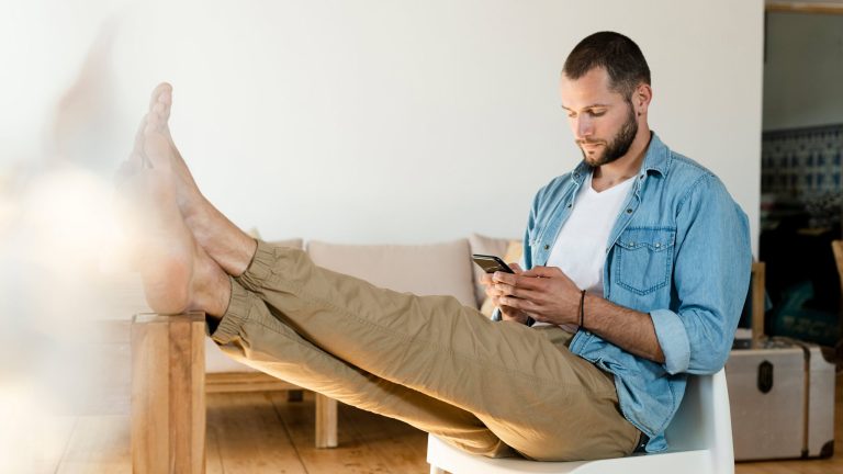 Eine Person sitzt auf einem Stuhl, hat die Füße auf dem Tisch und schaut auf ihr Smartphone.