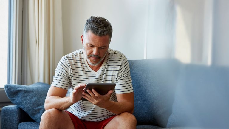 Eine Person sitzt auf einem Sofa und tippt auf ein Tablet in ihrer Hand.