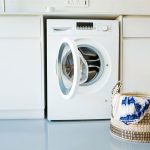 Eine geöffnete Waschmaschine steht unter einer Küchenzeile. Davor steht ein Wäschekorb, aus dem etwas Stoff hängt.