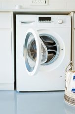 Eine geöffnete Waschmaschine steht unter einer Küchenzeile. Davor steht ein Wäschekorb, aus dem etwas Stoff hängt.
