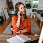 Eine Person sitzt an einem Tisch und h&auml;lt sich ein Smartphone ans Ohr. Vor ihr liegt ein Notizbuch.