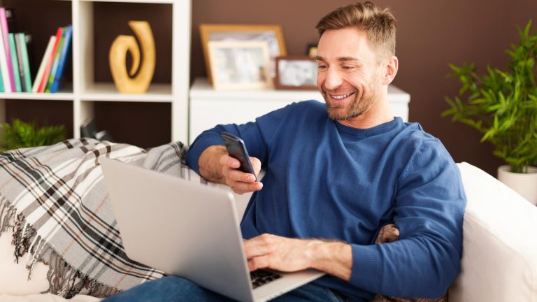 Eine Person sitzt auf einem Sofa mit einem Laptop auf dem Schoß. In der Hand hält sie ein Smartphone.