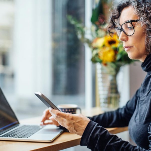 Eine Person sitzt vor einem Laptop. In der Hand h&auml;lt sie ein Smartphone, auf das sie schaut.