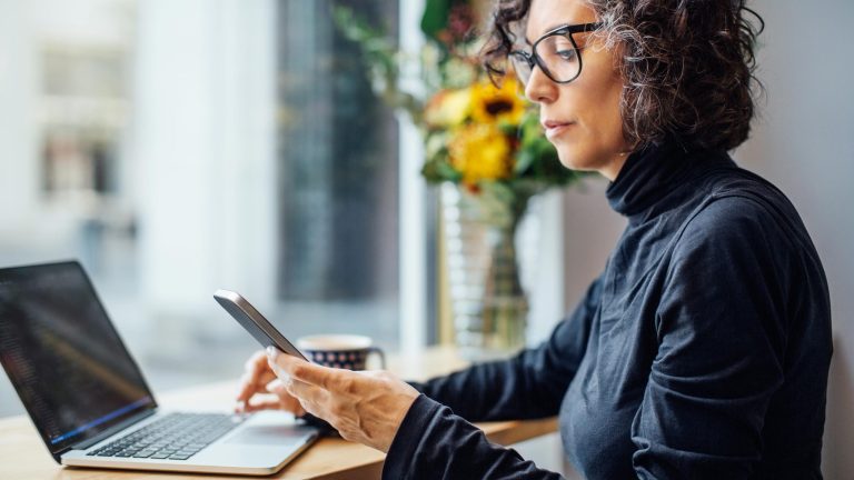 Eine Person sitzt vor einem Laptop. In der Hand hält sie ein Smartphone, auf das sie schaut.