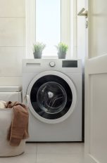 Eine Waschmaschine steht in einem Badezimmer. Daneben steht ein voller Wäschekorb.