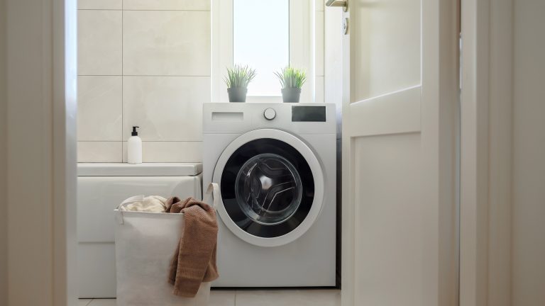 Eine Waschmaschine steht in einem Badezimmer. Daneben steht ein voller Wäschekorb.