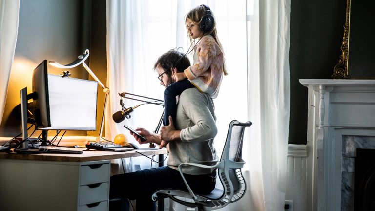 Eine Person sitzt vor einem Rechner, auf ihrer Schulter sitzt ein Kind mit Kopfhörern auf dem Kopf.